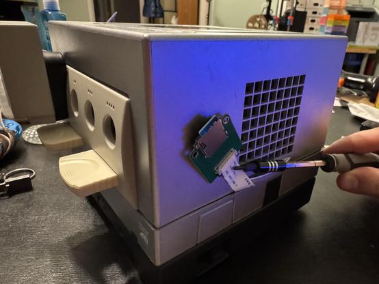 A picture of the Nintendo GameCube with a PCB hanging out the side with a ribbon cable going back into the GameCube