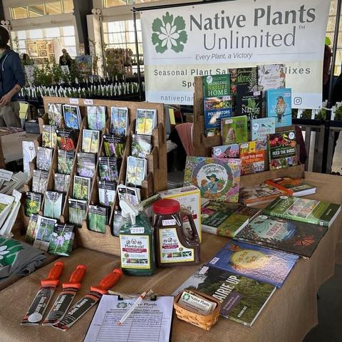 An event table with colorful products relating to native plants- books, seed packs, supplies. Sign in background reading, “Native Plants Unlimited- Every Plant A Victory”