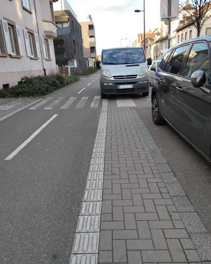 Un Opel Vivaro gris stationné sur le trottoir, entre les places de stationnements et la piste cyclable, rue Jacob. L’avant du véhicule occupe une partie du passage piéton qui traverse. Aucune visibilité sur la piste cyclable, la traversée piétonne ou la chaussée