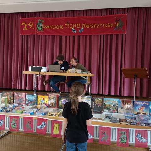 Ein Kind betrachtet die Auswahl der Preise für die besseren 50% des Turniers. Die Veranstalter sitzen auf der Bühne und werten aus.