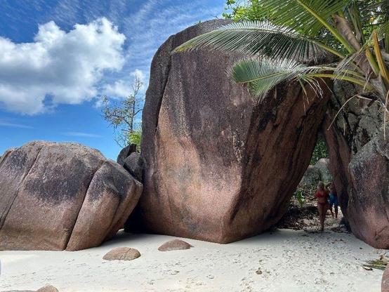 brown and dark brown giant rocks on a beach with almost white sand, clouds on the blue sky and palm trees, people walking underneath two rocks leaning on each other