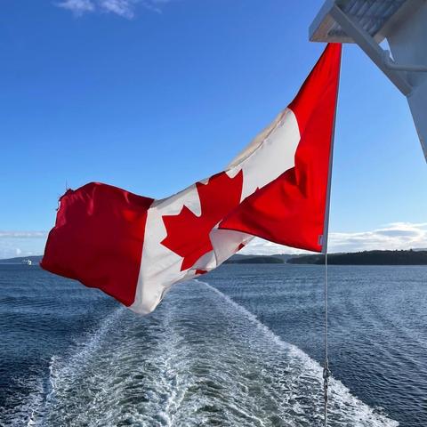 A Canadian flag waving in the wind, attached to a structure on a ferry. The flag is vibrant red and white, with the iconic red maple leaf in the centre. The background is a deep blue sky with a few clouds, as well as water with a visible wake from the vessel.