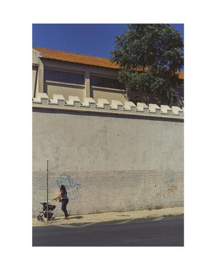 woman with babystroller walking in front of a big grey wall with graffiti, above a building and a tree