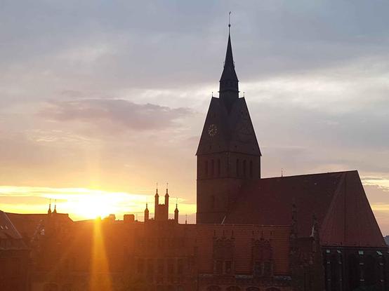 Eine Silhouette einer Kirche mit einem hohen Turm gegen einen Sonnenuntergang. Der Himmel zeigt Orange- und Grautöne, wobei das Sonnenlicht durch Wolken dringt. Im Hintergrund sind zusätzliche Gebäude sichtbar.