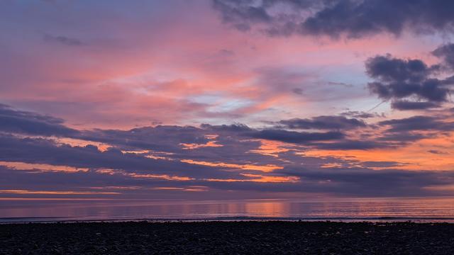 Historical Documentation in the form of a photograph of a rising sun over the ocean at Yirrell Beach in Winthrop, MA.  The sky is filled with vibrant colors of orange, pink, and purple, with the sun setting on the horizon and casting a warm glow over the water. The clouds are scattered across the sky, creating a dramatic contrast with the brightening sky as the day comes alive. The water is calm and reflects the colors of the sky. In the foreground, there is a rocky shoreline with small pebbles scattered across it. The overall mood of the image is peaceful and serene.
