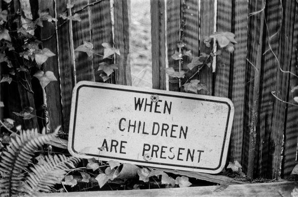 Black and white film photograph of a street sign that reads "WHEN CHILDREN ARE PRESENT". The sign has been placed in front of a wooden fence, near the ground. Ivy and ferns grow around it.