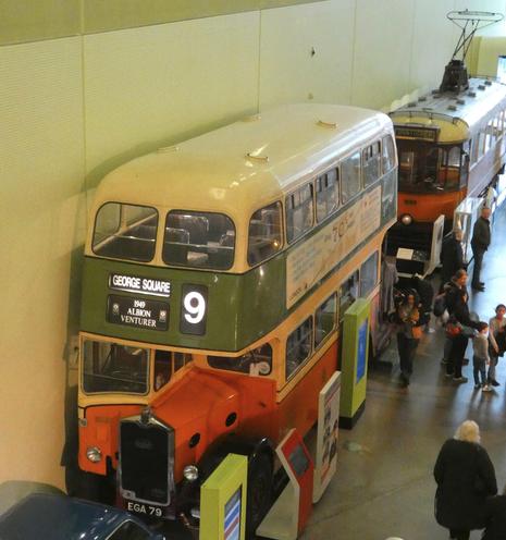 Viewed from above, a double decker bus parked against the wall, front facing us at an angle. Livery is cream/green/hellow. There is a vintage tram behind it and people walking by.