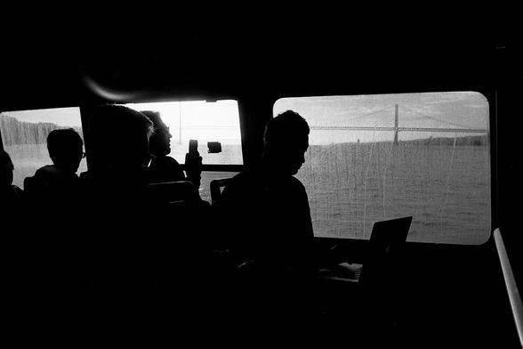 A black and white photograph captures the silhouettes of several people inside a ferry, with large windows showing a bridge over a body of water in the background. One person is using a laptop, while another holds up a mobile phone.