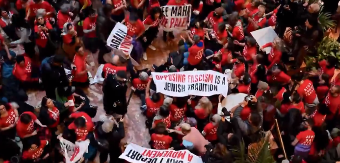 image from above of dozens of people sitting on the floor or standing next to each other, dressed in red & white tshirs and black pants and some of them holding banners. one of them clearly reads: opposing fascism is a jewish tradition.

on the front of some of the t-shirts it says, NOT IN OUR NAME. 

they are one of the many Jewish organizations protesting the abduction and imprisonment of Palestinian activist Khalil Mahmoud, by Trump and his fascist collaborators.