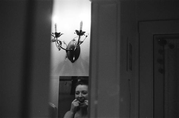 Black and white photograph of a young woman looking at herself in the mirror while flossing her teeth.