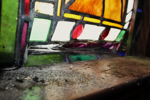 A windowsill covered in dust, debris and dead bees supports an aging stained glass window that is bowing and crumpling with age. Spiderwebs connect the warped window to the window sill.