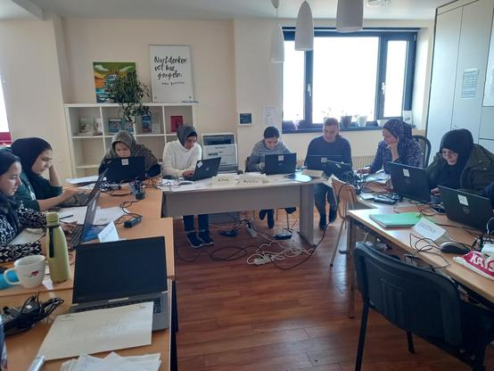 In einem Raum stehen mehrere Tische in U-Form, darauf knapp 10 aufgeklappte Laptops. Davor sitzen Frauen mit Migrationshintergrund und unterschiedlichen Alters. Sie erhalten von einer Lehrerin eine Einweisung und Schulung. 