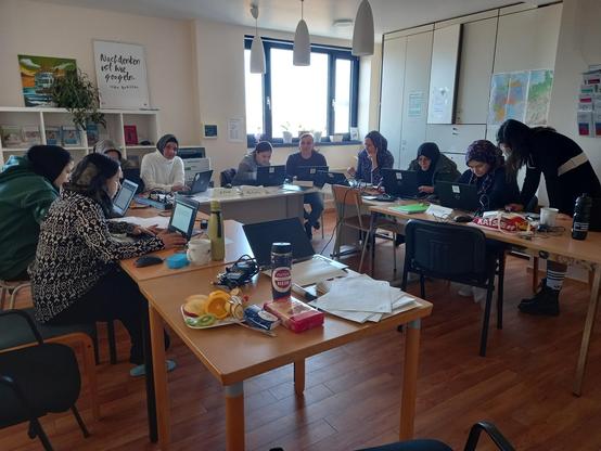 In einem Raum stehen mehrere Tische in U-Form, darauf knapp 10 aufgeklappte Laptops. Davor sitzen Frauen mit Migrationshintergrund und unterschiedlichen Alters. Sie erhalten von einer Lehrerin eine Einweisung und Schulung. 