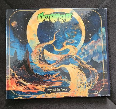 A CD album on dark background. It's very colourful and depicts a fantastic riverscape at night. There are multiple stars in the dark blue sky and two moons, the bigger of which is in the middle and forms an "O". There are dark rocky formations and a couple of trees to the left and right. Between them is a wide river in orange, red and yellow. The fluid in the river has bubble looking things embedded in it. A few tendrils of the liquid are floating up in the middle, curling into the sky through the big moon. The band name "Octoploid" is recognisable in it's light green logo under the upper edge, album name "Beyond the Aeons" is in a small polygonal panel above the lower edge.