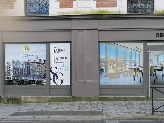 Photo de la façade d'une agence immobilière vantant des "prestations haut de gamme" avec un tag sur la vitre "tout est trop cher"