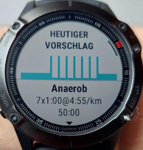 Man sieht eine Garmin Armbanduhr. Auf der Anzeige sieht man den Vorschlag für das heutige Lauftraining. Oben steht der Text "Heutiger Vorschlag", darunter sieht man ein Diagramm, das 7 Intervalle andeutet und darunter steht der Text "Anaerob, 7x1:00@4:55/km, 50:00".