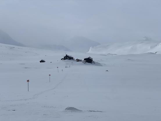 A couple black houses in the middle of a white plain. A clear backdrop just a bit behind them. Faint mountain tops at the distance. There’s no sky, just white dust. A skiing trail leading towards the photographer, marked with red crosses. 