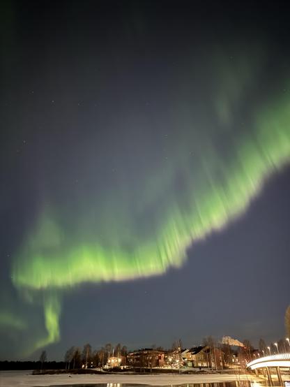 A bright green ribbon of the aurora snaking across the sky. Below is a frozen river and some low buildings. 