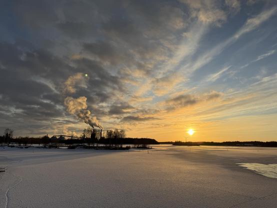 The low sun over a frozen river. There are several cloud layers merging with the smoke billowing from a distant building’s smoke stack. 