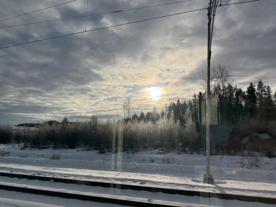 A shot from a train: the sun behind a sheet of clouds, illuminating a snow covered landscape with a dense pine-y forest. 