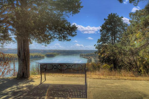 A wooden bench sits under the shade of tall trees, overlooking a tranquil Mississippi River below under a bright blue sky.