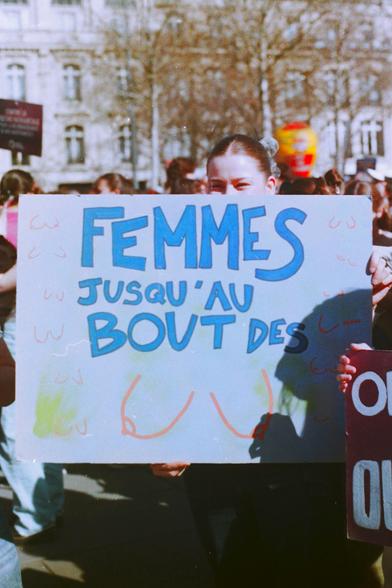 Une femme brandit une pancarte colorée lors d’une manifestation pour les droits des femmes. On peut lire en lettres bleues : "FEMMES JUSQU'AU BOUT DES", avec des dessins symbolisant la féminité autour du message. Derrière elle, une foule engagée marche sous un ciel clair, portant d'autres pancartes et revendications.