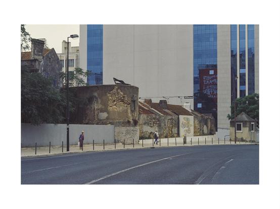 people walking on a sidewalk along historically old buildings in lisbon, all before a giant modern facade of glass and stone