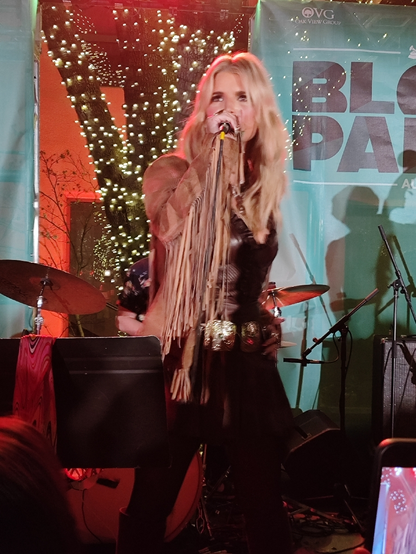 photo of Jessica Simpson, with long bleach blonde hair,  in a fringe tan leather jacket and brown leather top, singing into a microphone on a stage, with a tree with white lights, and banners with "Block Party" written on them, in the background