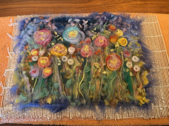 Multicoloured bits of wool roving laid out in a soft floral composition. Rectangle shaped on bubble wrap and bamboo mat, all on top of wooden surface. Ready for wet felting!