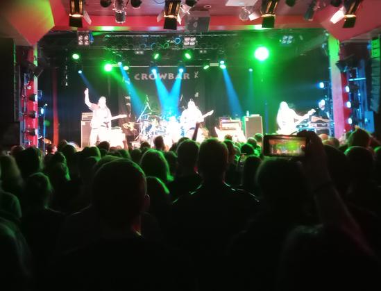 Colorfully lighted stage, seen from dark auditory, four musicians (git, git/voc, b, dr) performing.