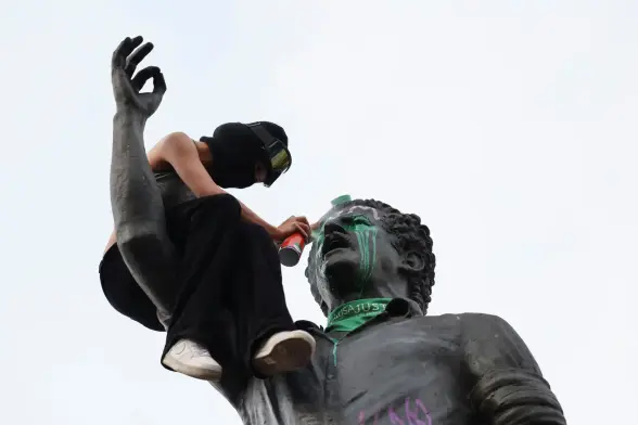 A young woman sprays paint on the statue of Luis Carlos Galán wearing a green scarf reading "Just Cause," sitting on his raised arm.