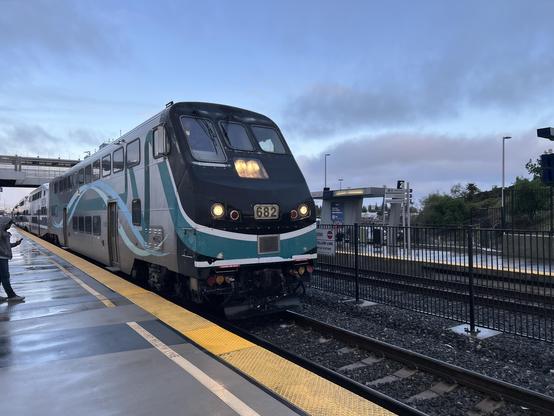 The front of a Metrolink train, a double-deck silver and dark blue passenger train, pulling into Anaheim Station. The sky is cloudy and the ground is very wet.