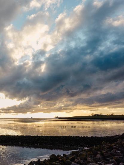 The photo captures a dramatic sky filled with dark, heavy clouds mixed with lighter, fluffy ones. The sun is partially hidden behind the clouds, casting a golden light that reflects off the calm water below. The contrast between the dark clouds and the bright patches of sky creates a striking and moody atmosphere.