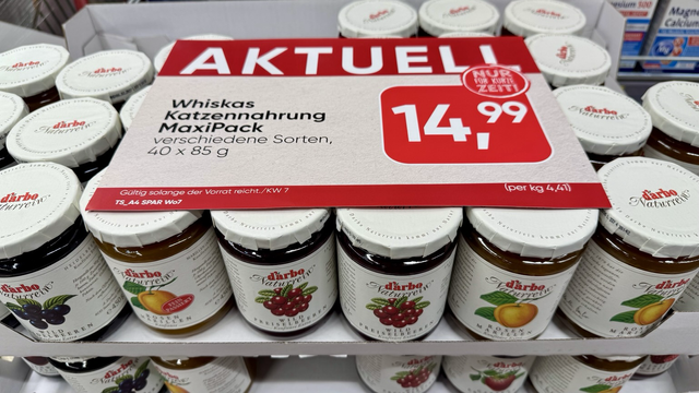 Im Supermarkt sind Gläser mit Konfitüre übereinandergestapelt: Heidelbeeren, Marillen, Erdbeeren, Preiselbeeren und andere. Oben drauf liegt ein Schild: "AKTUELL: Whiskas Katzennahrung MaxiPack, verschiedene Sorten".
