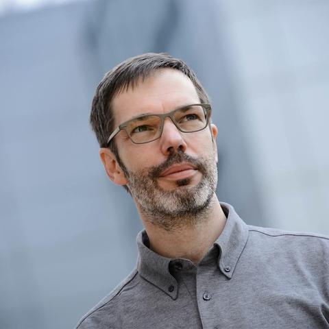 Portrait von DJ Franksen. Er trägt eine Brille, ein graues Hemd und einen Drei-Tage-Bart und steht vor einem verschwommenem grauen Hintergrund.