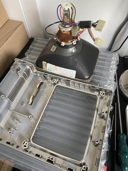 Picture of the front panel of a Macintosh Plus laying disassembled in front of the CRT of the computer, both laying on top of plastic bubble wrapping, used to protect them and the desk they’re laying on, with some screwdrivers by the side.