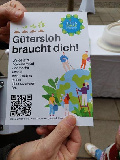 Person hält einen Flyer mit Abbildungen von Garten- und Malern. Der Text lautet: „Gütersloh braucht dich!“ mit einem QR-Code und einer Webadresse für weitere Informationen.