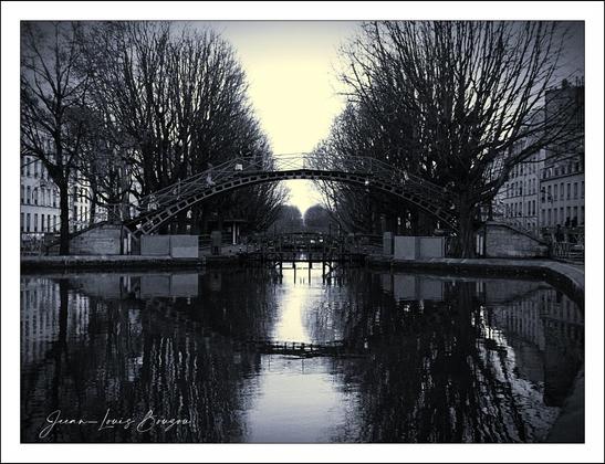 The image presents a serene scene with a gentle, almost ethereal quality. At the center, a delicate arch bridge gracefully spans a tranquil canal. The reflections on the water create a mirror-like effect, enhancing the symmetrical beauty of the setting.
Flanking the canal, tall trees, stripped of their leaves, stand like silent sentinels, adding a sense of stillness and contemplation to the atmosphere. The monochromatic palette amplifies the mood, evoking a timeless essence that feels both nostalgic and poignant.