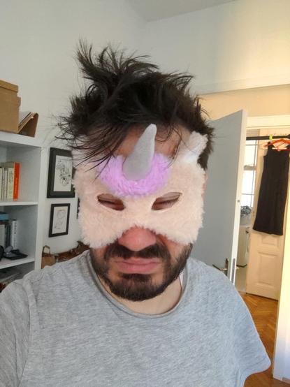 man wearing a too tight unicorn mask, covering half of his face, making his eyes look squeezed shut. He has crazy hair and wears a grey t-shirt