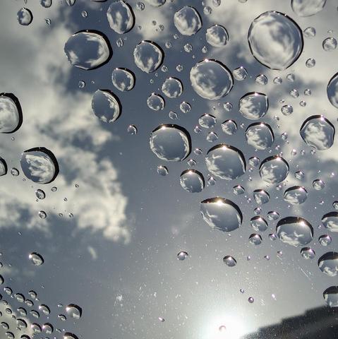Rain drops on the sunroof of a car looking up toward the blue sky with big white puffy clouds