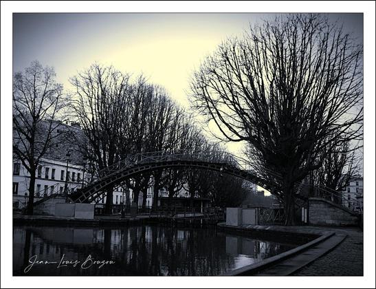 The image captures a serene, almost haunting scene. A graceful bridge arches elegantly over a still body of water, reflecting its curvature in the dark surface below. The landscape is framed by towering trees, their bare branches reaching skyward, evoking a sense of stark beauty in the absence of leaves.
Soft contrasts between light and shadow create a gentle ambiance, enhancing the calmness of the setting. The subdued tones suggest an atmosphere of tranquility, perhaps hinting at the passage of time or the quietude of a winter's day.
A nearby structure peeks through the trees, its architecture softened by the monochrome palette, blending into the serene backdrop. This image may symbolize a moment of pause, inviting viewers to reflect and contemplate, perhaps evoking feelings of nostalgia or peaceful solitude. The stillness of the water serves as a mirror, touching on themes of introspection and the connection between nature and human presence.