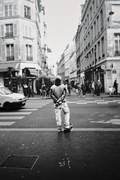 Un homme, debout sur son skateboard au milieu d'un passage piéton parisien, mains croisées derrière le dos, observe tranquillement la rue animée devant lui. La scène urbaine est capturée en noir et blanc, accentuant l'atmosphère contemplative et intemporelle.