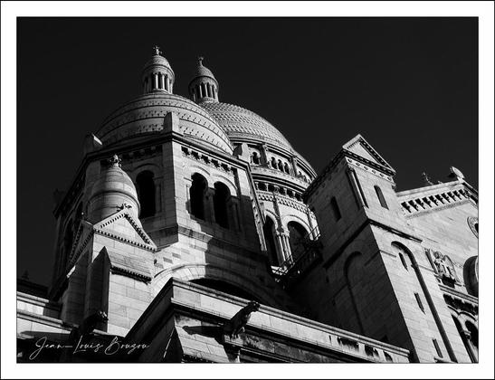 The image captures the silhouette of an architectural marvel, with its grand domes reaching towards the sky. The structure, characterized by its intricate stonework, appears majestic against a stark black background that enhances its features. The play of light and shadow brings out the textured details of the building, emphasizing the craftsmanship that has gone into its design.
The domes are prominent, their rounded shapes contrasting with the straight lines of the surrounding walls. This juxtaposition creates a sense of movement and harmony, hinting at both strength and elegance. The absence of color transforms the scene into a timeless rendering, inviting the viewer to appreciate the beauty found in form and structure alone.
One might interpret this image as a symbol of aspiration, with the dome representing a connection to the heavens. The perspective from below lends a feeling of grandeur, as if the building is towering over its surroundings, evoking feelings of admiration and wonder. It stands as a testament to human creativity and perseverance in architecture, inspiring inspiration and contemplation.