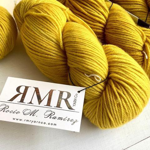 Angled view of several bright yellow skeins of yarn on a white plank background. There is a white label attached, with RMR on it in shiny letters.
