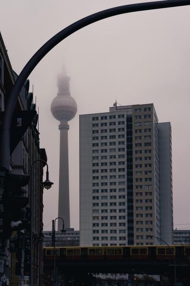 A photograph of an urban scene in Berlin on a foggy day. In the foreground, a yellow-red S-Bahn train crosses a bridge. Behind it, a large residential building with many windows stands tall. In the background, the Berlin TV Tower rises into the sky, with its top disappearing into the dense fog. A curved streetlamp and various city elements frame the image.
