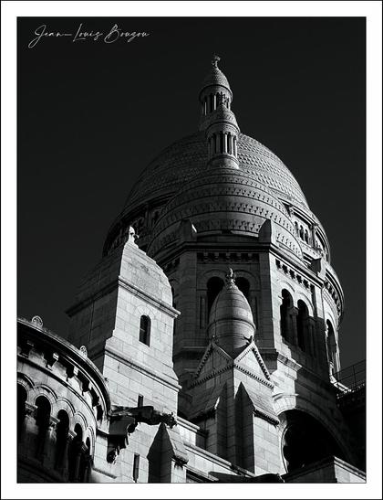 The image captures the impressive architectural structure of the Sacré-Coeur basilica, with a focus on its grand dome and intricate details. The building, rendered in striking black and white, emphasizes the textures of the stone and the delicate contours of the dome's curves. The play of light and shadow enhances the sense of depth, highlighting features like the ornate turrets and the layered façade.
This choice of monochrome adds a timeless quality, drawing attention to the architectural design rather than the colors that might distract. The overall composition evokes a sense of majesty and tranquility, inviting viewers to reflect on the building's presence. The dome itself, reaching upward, suggests aspirations and a connection to something greater, perhaps representing faith, community, or artistic achievement. The image as a whole can be seen as a tribute to the craftsmanship and vision behind such an enduring structure.