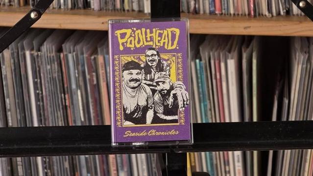 Das Bild zeigt eine Kassette mit dem Titel "PooLhead: Seaside Chronicles", die prominent dargestellt ist. Das Cover des Bandes hat ein farbenfrohes Design mit drei illustrierten Personen, die vor einem Hintergrund aus gestapelten Platten stehen.
