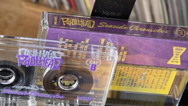 Das Bild zeigt eine Kassette mit dem Titel "Seaside Chronicles" von Poolhead. Die Kassette befindet sich in einem durchsichtigen Gehäuse mit schwarzen Bandrollen und lila Akzenten. Die Vorderseite des Gehäuses enthält den Namen des Künstlers und den Albumtitel in stilisiertem Text.
