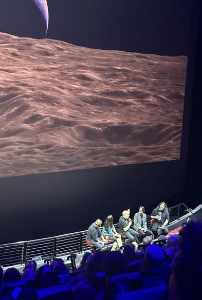 A row of five people on chairs at the front of a large IMAX audience, with a picture the Moon and Earthrise on the screen behind. 