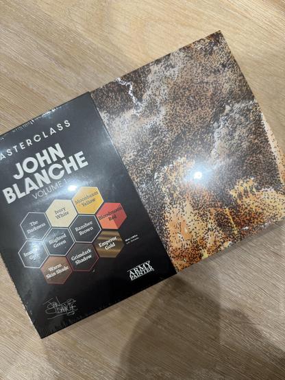 A boxed set titled "Masterclass John Blanche Volume 1" featuring various paint colors with names listed in hexagonal shapes, alongside an abstract textured design on the right. The box is partially opened and placed on a wooden surface.